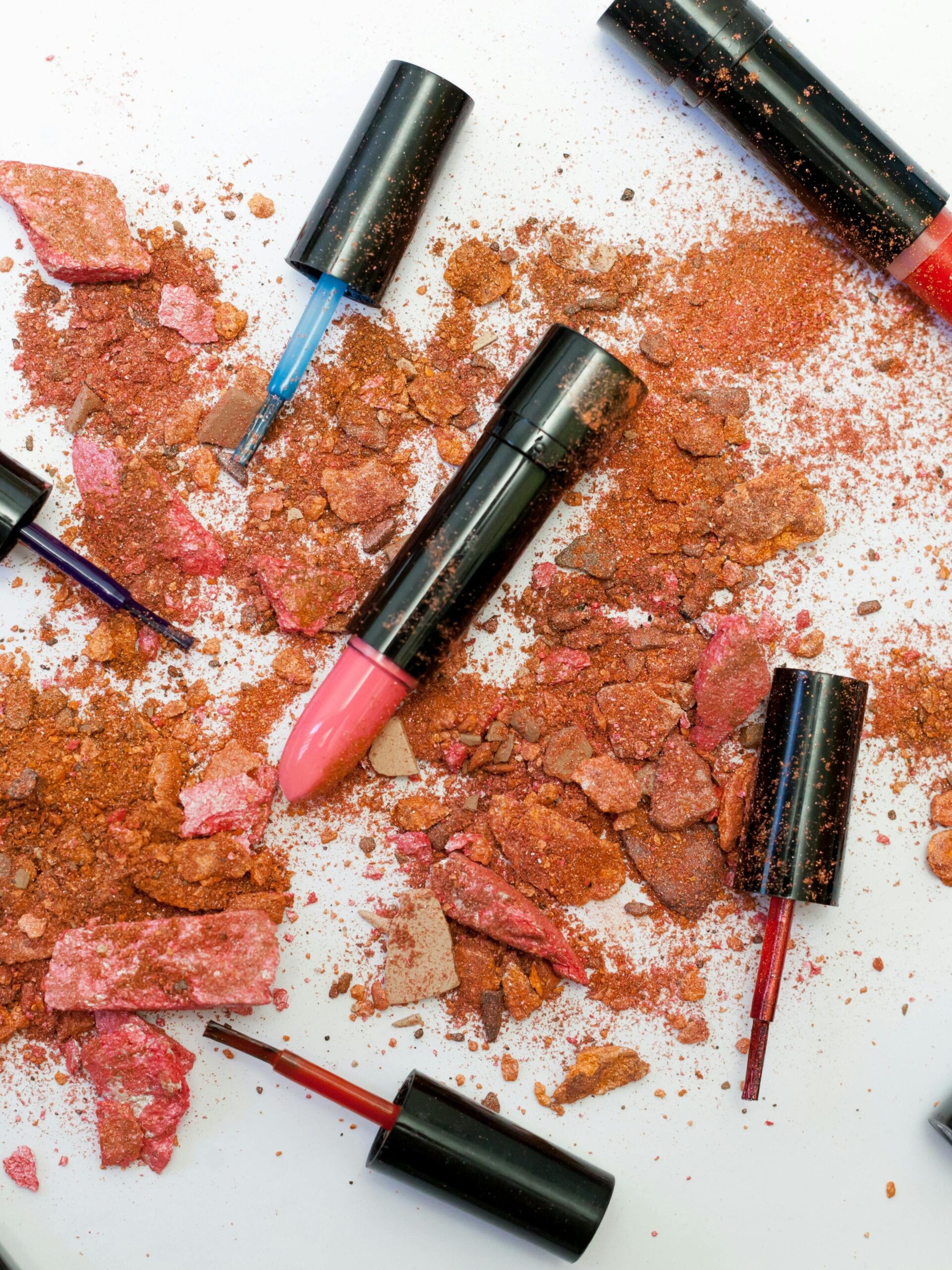 Home Colorful scattered makeup products including lipsticks and crushed powders on a white background.