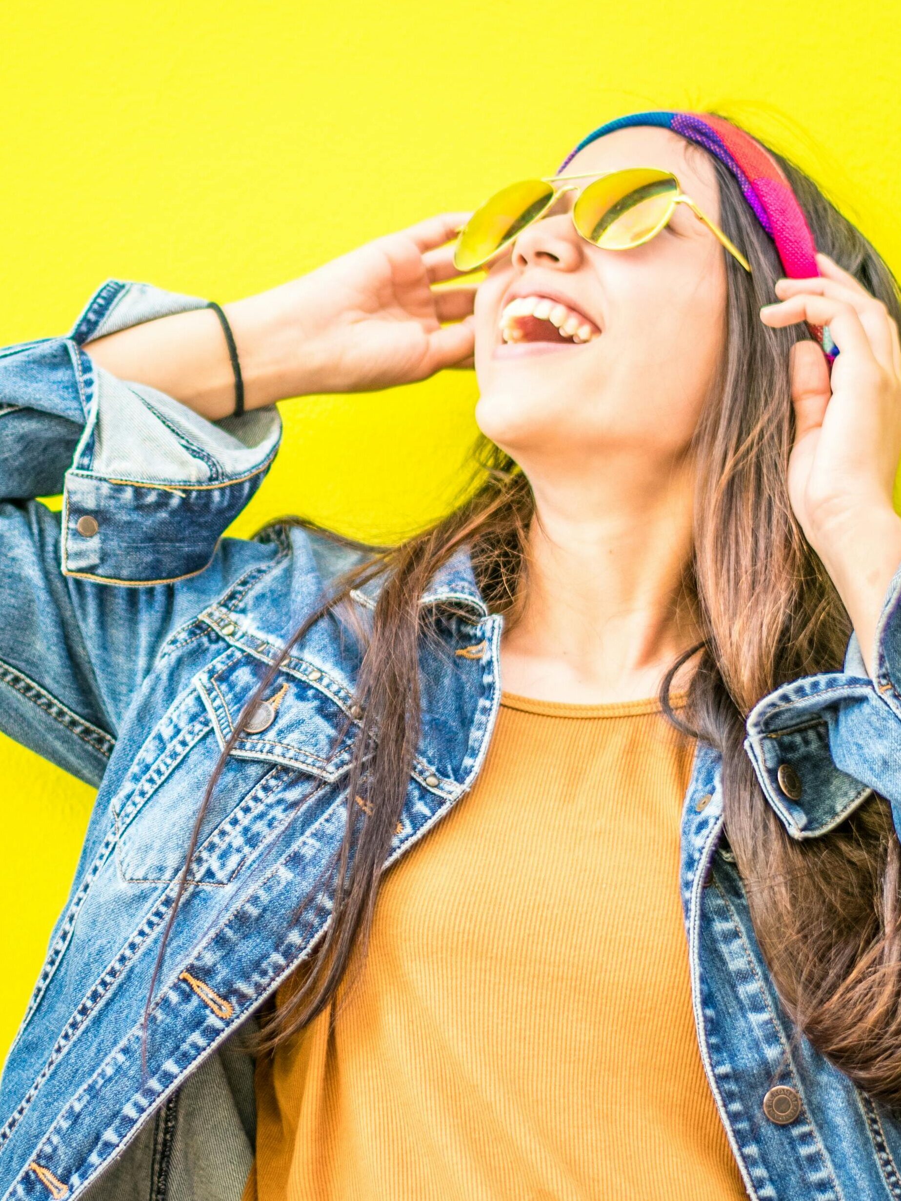 Home Smiling woman in sunglasses stands against vibrant yellow wall, radiating happiness.